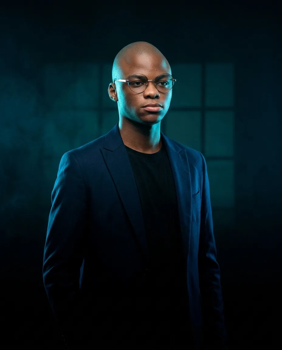 Yoane Ngassam, portrait en veste sombre, lumière basse, fond uni.